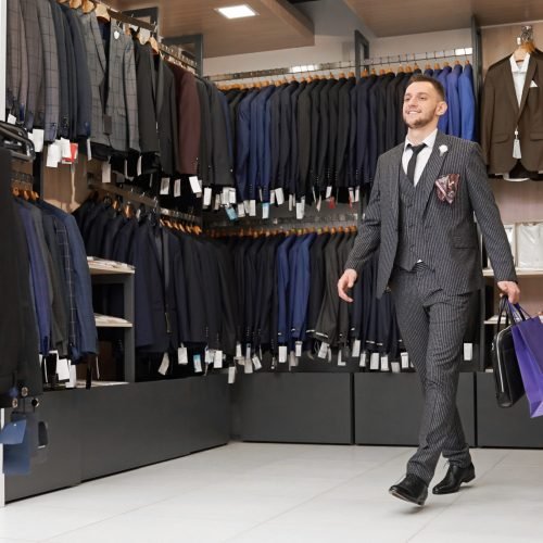 Happy client in striped suit, white shirt, necktie in boutique with shopping bags. Elegant gentleman with beard doing shopping in store. Fashionable clothing and shoes for men.